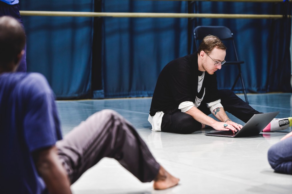 Rob sits on the floor of a dance studio, working on his laptop